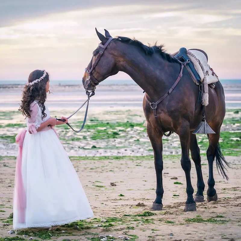 Niña de comunión llevando un caballo por la playa al atardecer